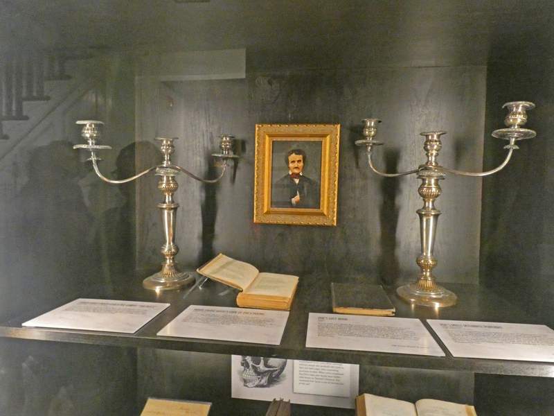 A apir of candlesticks, some papers and books with Edgar Allan Poe's Portrait on wall.