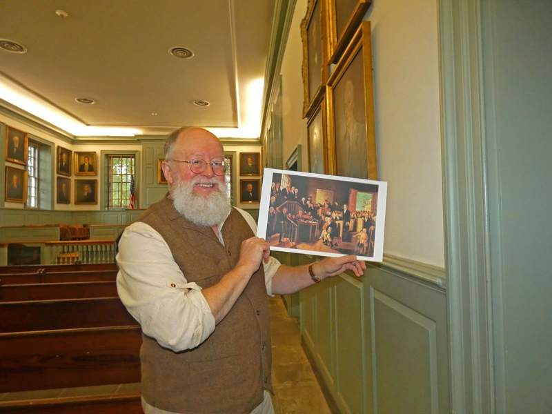 A man in Colonial dress holds a picture of a courthouse with people dressed i Colonial garb.