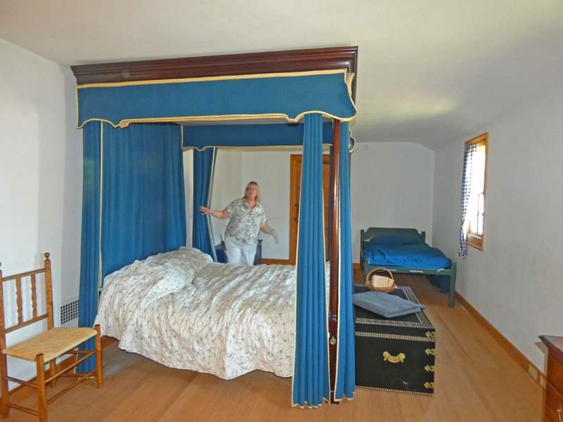 A woman standing behind a colonial style four poster bed with blue drapes.