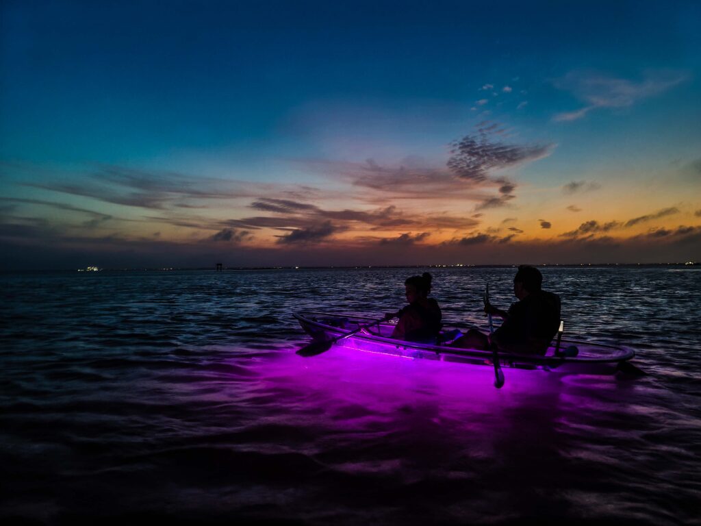 Two people in a kayak glowing purple in the water at dusk.