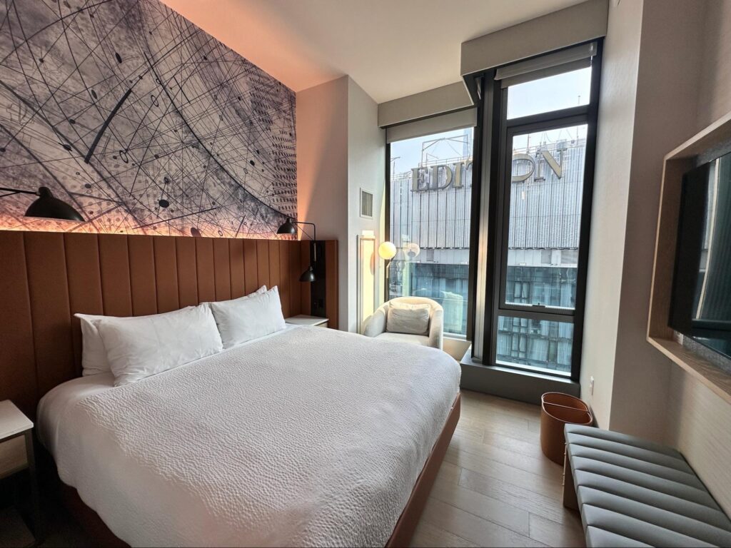 Standard King room at the Tempo Hilton Times Square with white bedding, a gray bench and tv on the opposite wall, a gray line drawing above the bed and white chair in front of the floor-to-ceiling-windows.