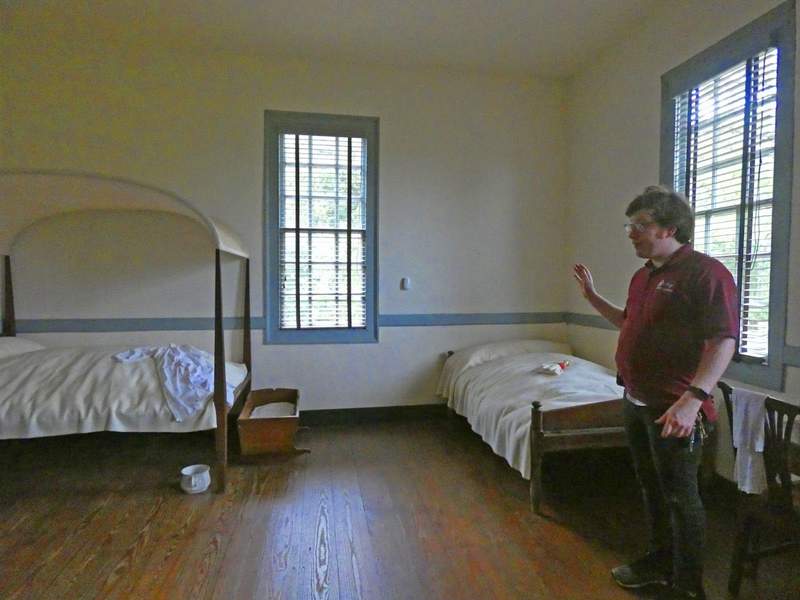 Man standing in a colonial era bedroom with two beds and a chamberpot.