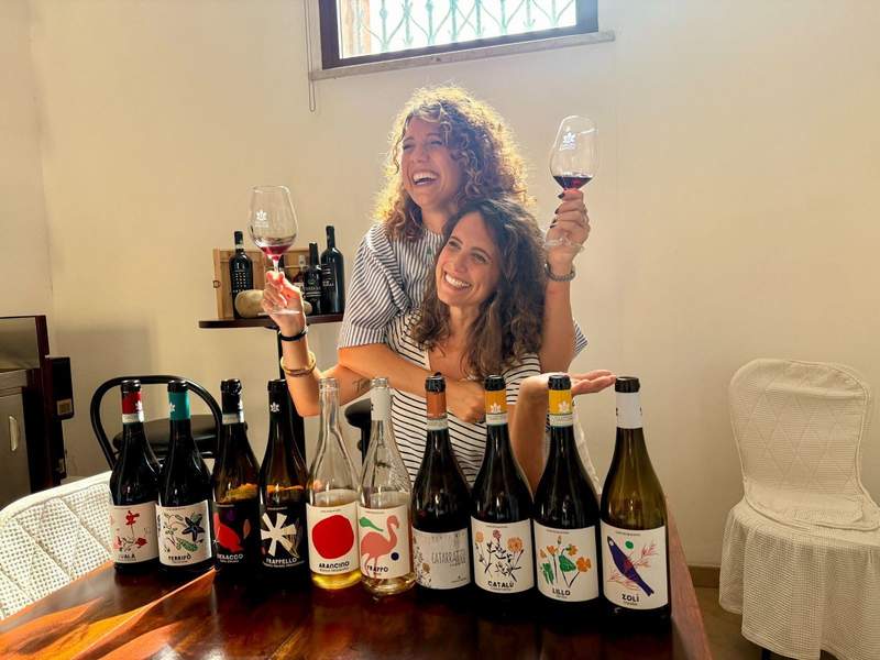 Sisters Rosanna and Giovanna Minini of Caruso & Minini winery smile with wine glasses in hand over a selection of wine bottles.