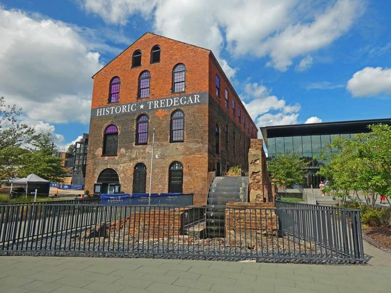 There story brick building with a water wheel in front and modern glass building on right side.