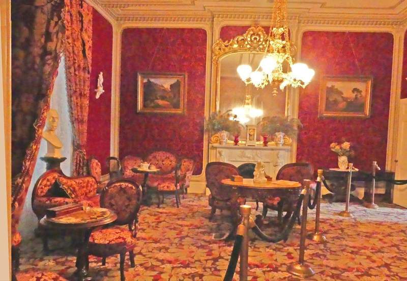 Victorian style room with red wall paper and red upholstered sofa and chairs with white fireplace and mirror and pictures on wall.
