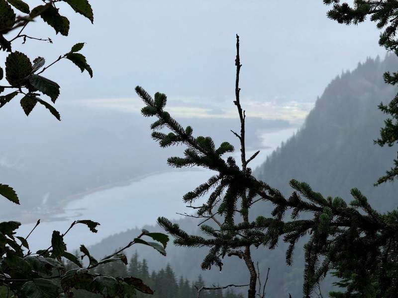 View from the top of a mountain looking down on the town of Juneau and a channel. Mountains and clouds in the background.