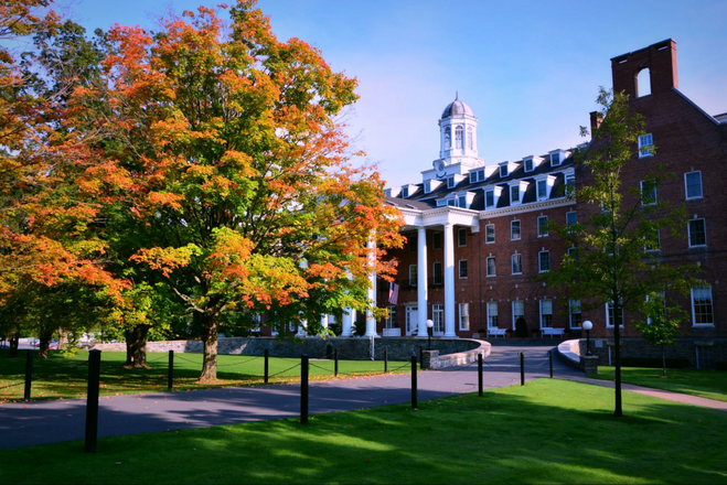 The entrance to The Otesaga Resort Hotel, as seen in fall. The building is red brick and ornate, with tall white columns that frame its porte cochere. A tree with leaves of orange and yellow is seen in the foreground.