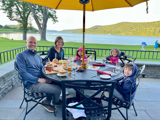 A family eats at a patio table at The Otesaga Resort Hotel's Hawkeye Bar & Grill in Upstate New York, featuring scenic seating overlooking a pristine lake.