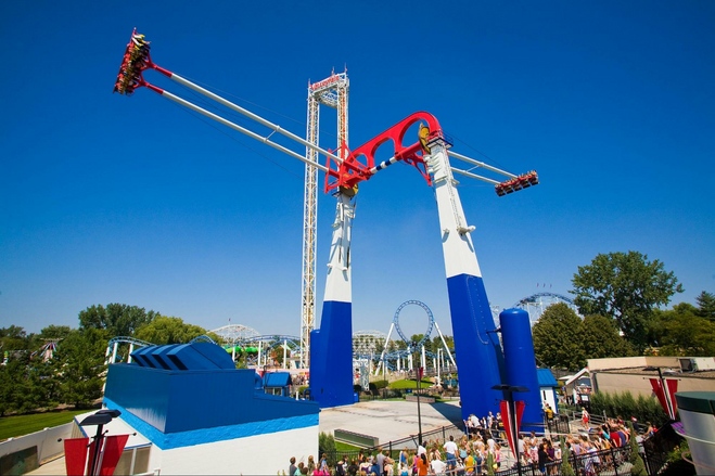 A red, white and blue amusement park attraction against a clear blue sky. People are sitting in seats and swining high in the air on either side.