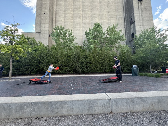 Two boys playing corn hole. One boy is wearing black. The other, younger boy is in a grey sweatshirt holding a red bucket.