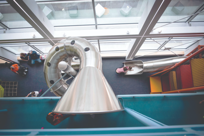 An overhead shot shows a big silver funnel goes into a blue container. There is a slide with a toddler attempting to climb up, but they are being stopped by a parent. Padded yellow and orange steps lead to the slide.
