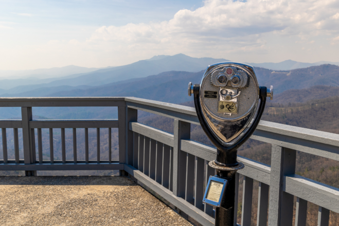 A silver coin-op binocular device next to a grey railing overlooking hazy mountains off in the distance from a lookout point in Blowing Rock, North Carolina.