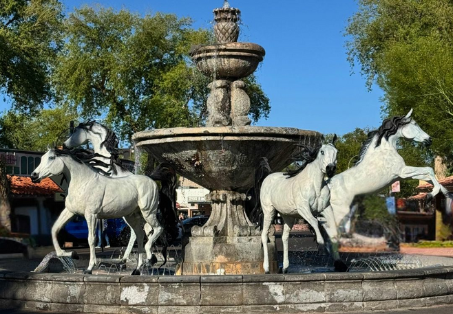 Stone fountain with white horse statues in the center of Old Town Scottsdale surrounded by trees and adobe-style buildings