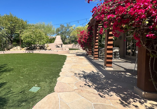 Pathway lined with stone pavers and vibrant pink bougainvillea at Four Seasons Scottsdale at Troon North, with green grass and a desert-inspired fireplace in the background.