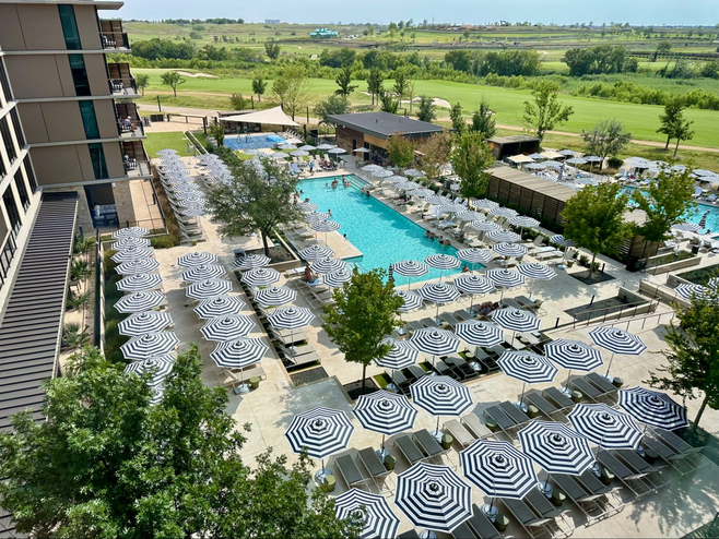 Aerial view of the main outdoor pool at the Omni PGA Frisco Resort, an excellent Frisco sports getaway hotel, lined with black and white striped umbrellas with golf course holes visible in the background.