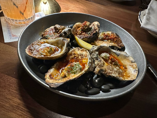 Broiled oysters and lemon wedge served atop black pebbles on a wooden table at Trick Rider in Frisco, Texas.