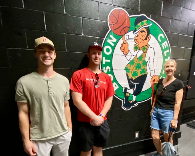 Two young guys and their mom standing against a black wall next to the Boston Celtics logo.