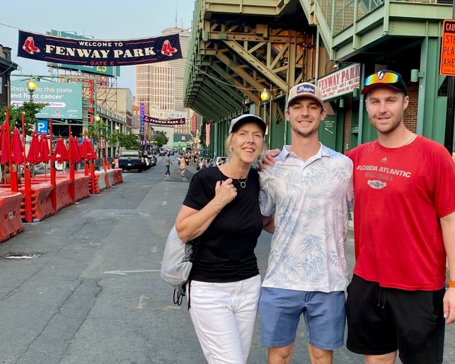 A lady in white jeans with her two young adult sons in Boston Fenway Park.