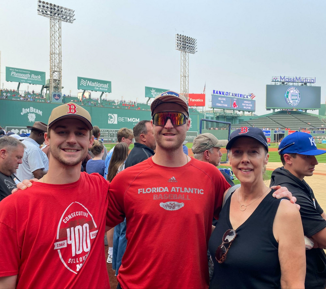 Two young adult guys in red shirts standing next to their mom in Fenway Park.