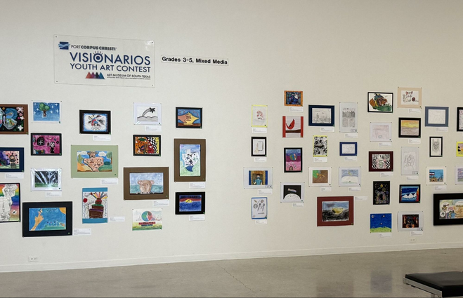 Various drawings and paintings by local South Texas schoolchildren displayed on a wall at the Art Museum of South Texas, with a sign stating that these are part of the Visionarios Youth Art Contest, Grades 3-5, Mixed Media.