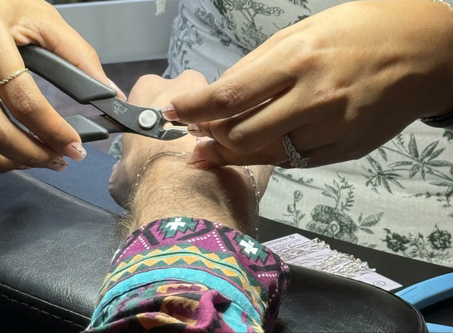 A pair of hands holds a wire cutter, measuring and cutting a silver chain around the wrist of a gentleman in a brightly-colored, long-sleeved shirt.