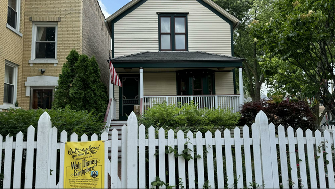 A yellow corner home with a white picket fence next to a large tree and an American flag.