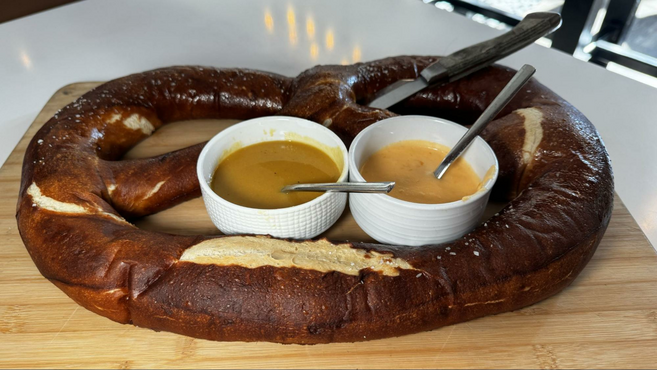 A gigantic two-pound pretzel with two circular cups filled with beer cheese and honey mustard on a wooden board.