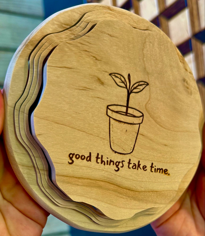 Carved into the wooden base of a basket ready to be put together is a simple flower pot holding a stalk with two leaves and the words in lowercase: good things take time.