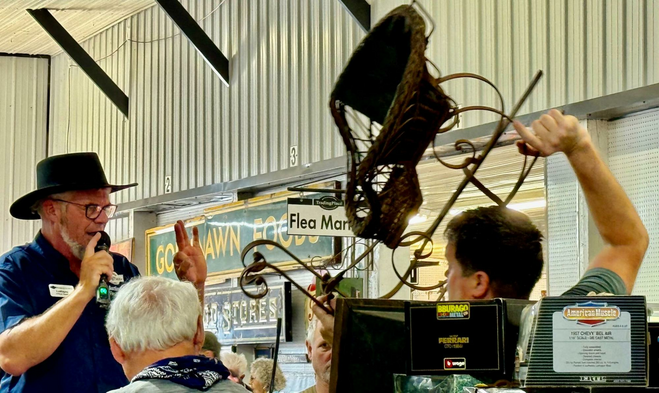 Auctioneer wearing a broad-brimmed black Amish hat holds up his left-hand pinky, ring and middle fingers while his right hand holds a microphone. Helper hoists a child-sized sleigh over his head so shoppers can call out their bids.