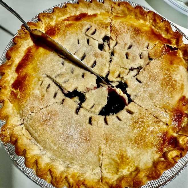 A hint of cooked blueberry seeps through the crust where a metal pie server has sliced triangle shaped pieces. The pie crust is a blend of beige dusted with sugar and dark crisp curved edges with the silver pan edges circling the crust.