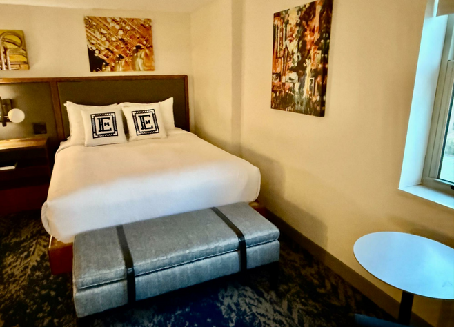 Queen-size bed with two pillows bearing a big letter "E" for Elkhart has a grey bench at the foot, the width of the bed. Walls are a calm yellow-beige.