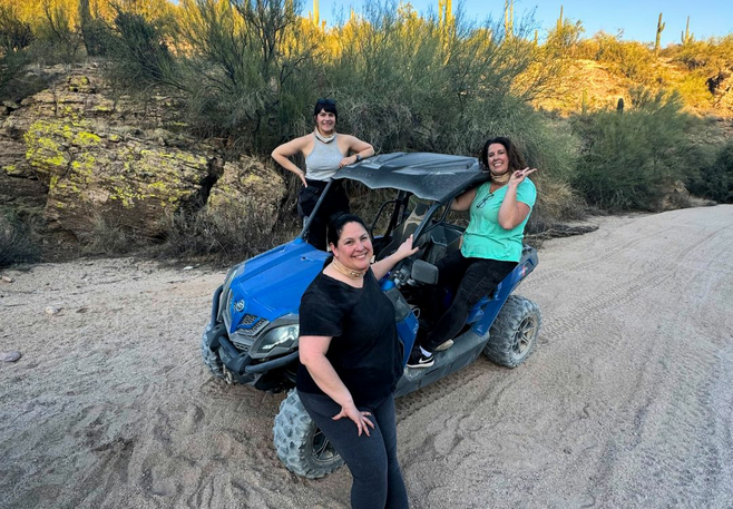 3 women pose on a dune buggy in the sand with desert flora behind them.