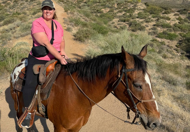 A woman in a pink shirt and ball cap sits atop a brown horse in the desert.