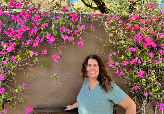 A woman in a green shirt poses smiling surrounded by pink flowers