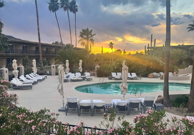 the sunset with palm trees and a cloudy sky with a pool and sunchairs surrounding it.