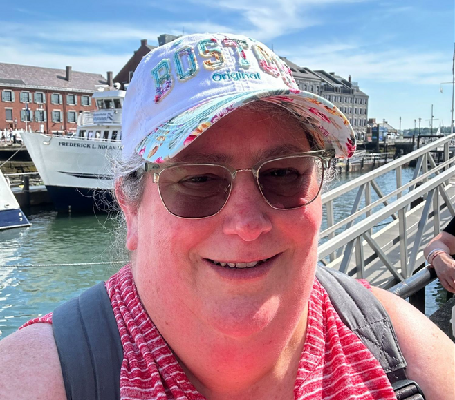 Woman poses in front of Boston harbor. She is wearing a white and floral Boston cap, dark tinted glasses, a red and white striped tank top and gray backpack straps.