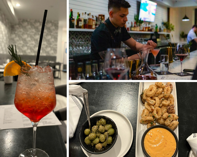 A bartender crafting a cocktail at the bar.A wine glass with red liquid at the bottom and clear liquid at the top with a orange garnish and rosemary sprig.Small plates of food including a bowl of olives, calamari with a sauce on the side, and focaccia slices.