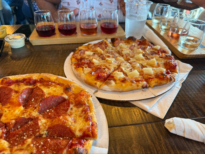 Two pizzas in the foreground, one Hawaiian and one pepperoni, with 2 wine flights of 4 wines each in the background.