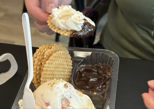A hand holds a round waffle chip dipped in custard and hot fudge, over a plastic container with more waffle chips, custard and hot fudge and a spoon sticking up.
